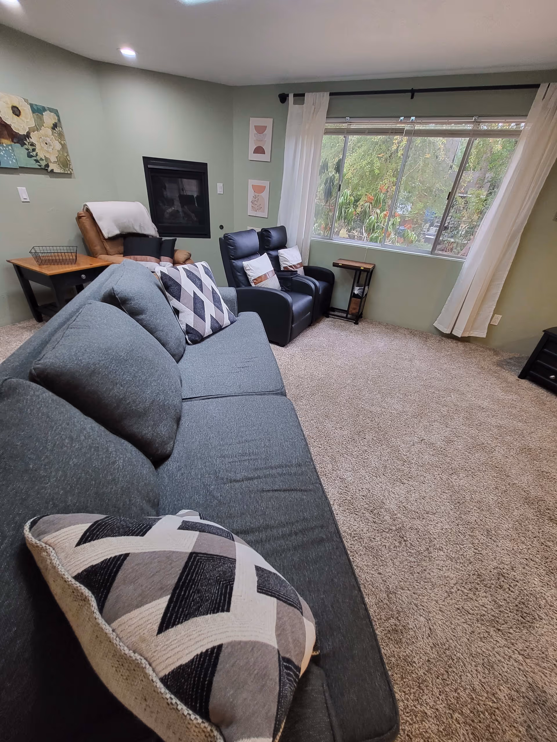 A cozy living room with a gray couch adorned with patterned pillows in the foreground. Behind the couch are two black recliners with white and brown pillows, and a brown armchair with a white throw blanket. The walls are painted light green, decorated with floral and abstract artwork. A large window with white curtains lets in natural light and shows greenery outside. The floor is carpeted in beige.
