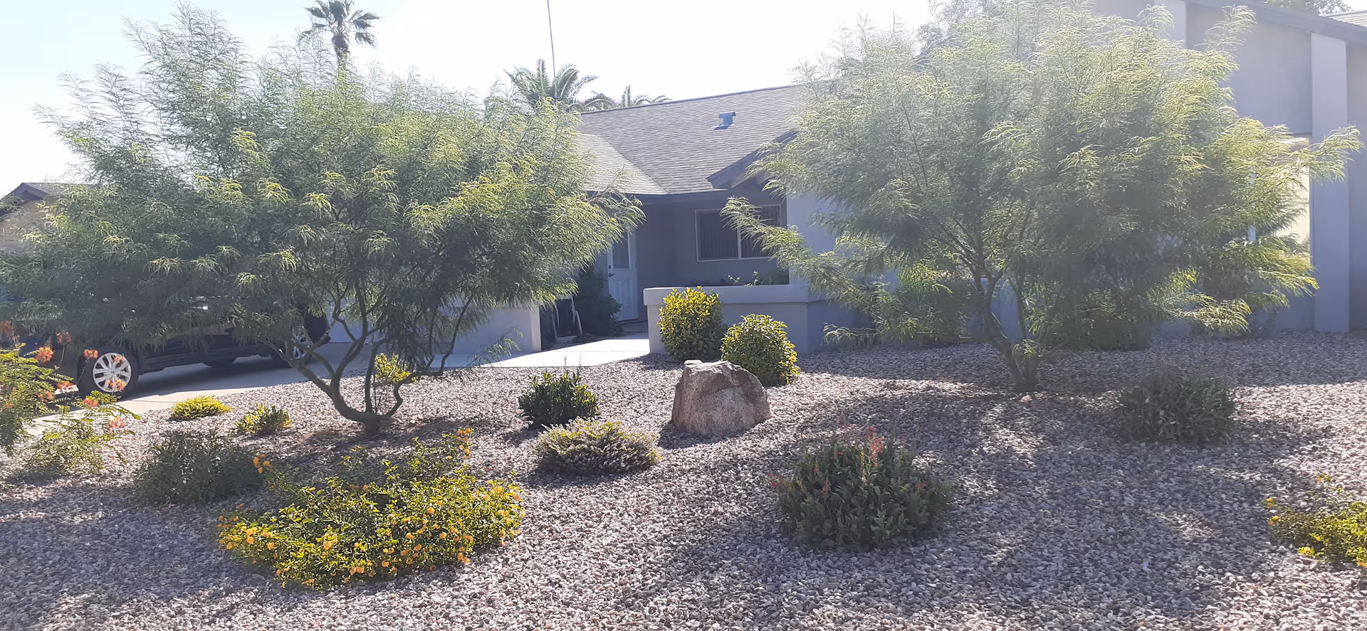Front view of a single-story building with a gravel yard featuring various shrubs and small trees. The building has a covered porch area and a dark roof, with a car partially visible on the left side.