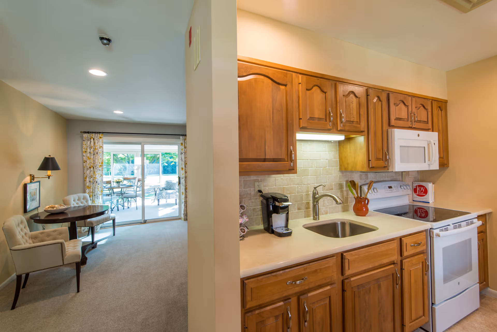Interior view showing a kitchen area with wooden cabinets, a white stove, microwave, and sink. Adjacent to the kitchen is a small dining area with two upholstered chairs and a round wooden table. Sliding glass doors lead to an outdoor patio with additional seating.