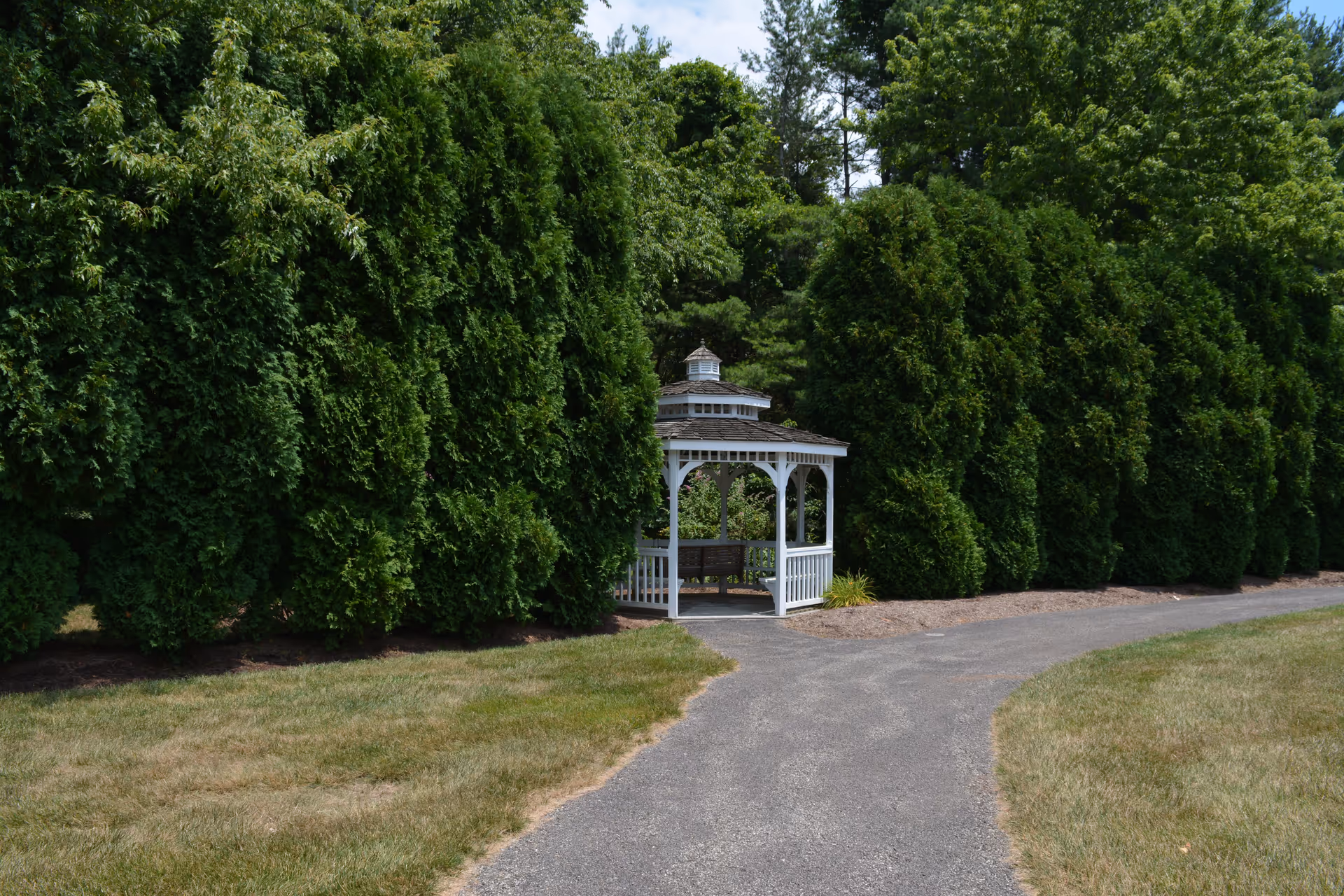 A white wooden gazebo with a bench inside, situated along a paved pathway surrounded by tall green trees and grass in an outdoor garden area.