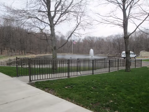 Outdoor scene showing a fenced pond with a water fountain in the center, an American flag on a pole near the pond, leafless trees surrounding the area, a paved sidewalk, green grass, a white van parked near a small shed, and a cloudy sky.