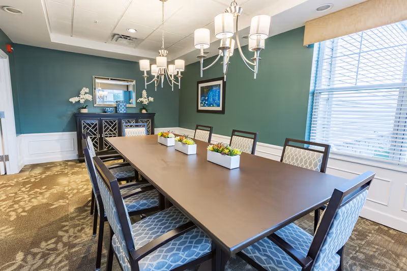 A well-lit dining room with a long rectangular wooden table surrounded by eight cushioned chairs with patterned upholstery. Three small rectangular planters with green and yellow succulents are placed on the table. The walls are painted green with white wainscoting, and there is a sideboard with decorative items and a mirror above it. Two modern chandeliers hang from the ceiling, and a large window with blinds allows natural light to enter the room.