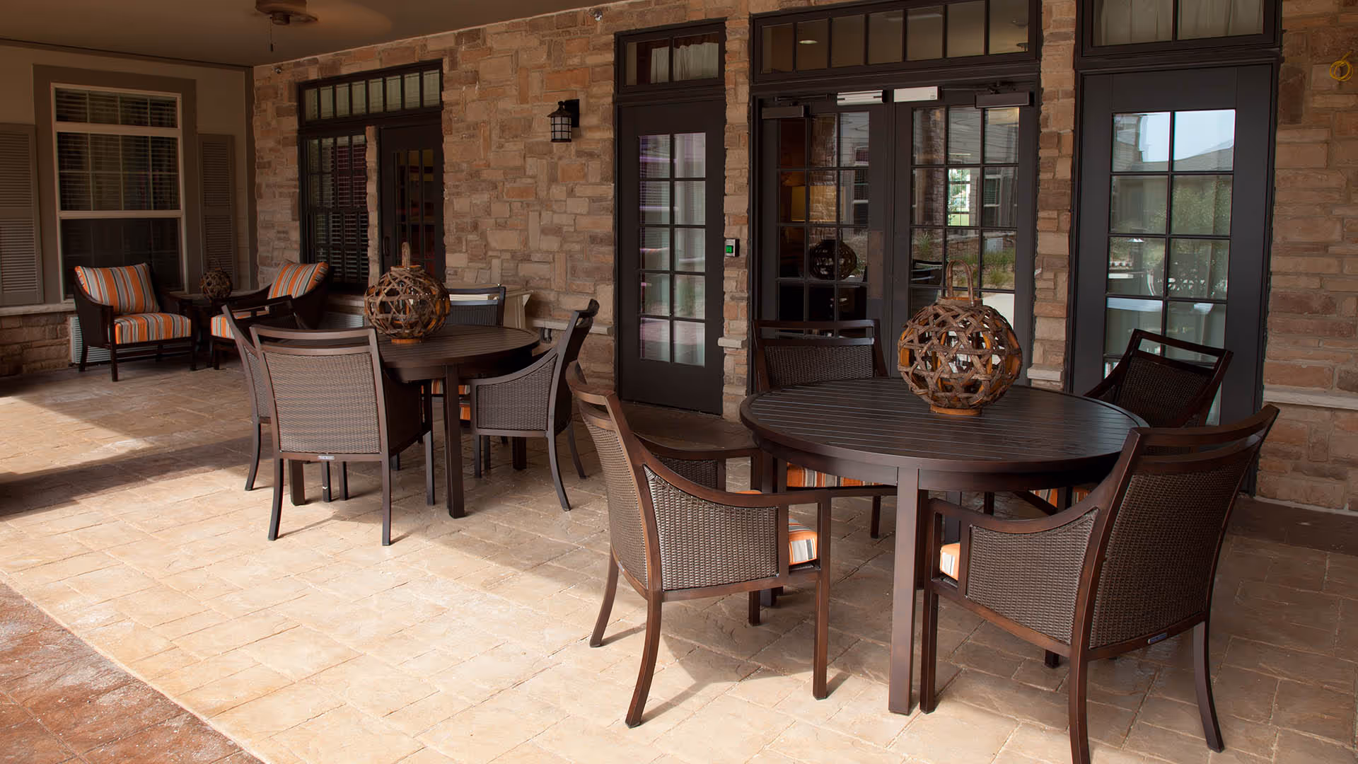 Covered outdoor patio with round wooden tables and wicker chairs arranged in front of a stone-clad building.