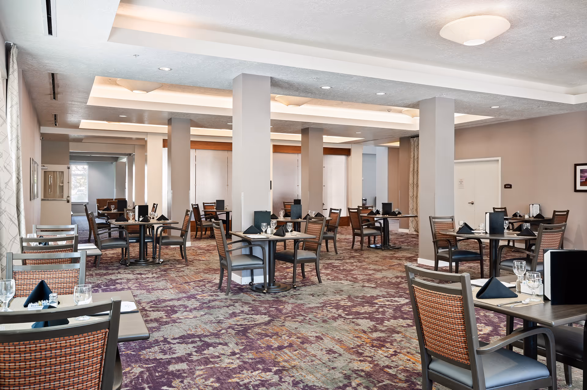 A spacious dining room with multiple tables and chairs arranged neatly. Each table is set with black napkins, glasses, and menus. The room features a patterned carpet in shades of purple and beige, light-colored walls, and ceiling lights. Several pillars are present throughout the room, and there are large windows with curtains on one side.