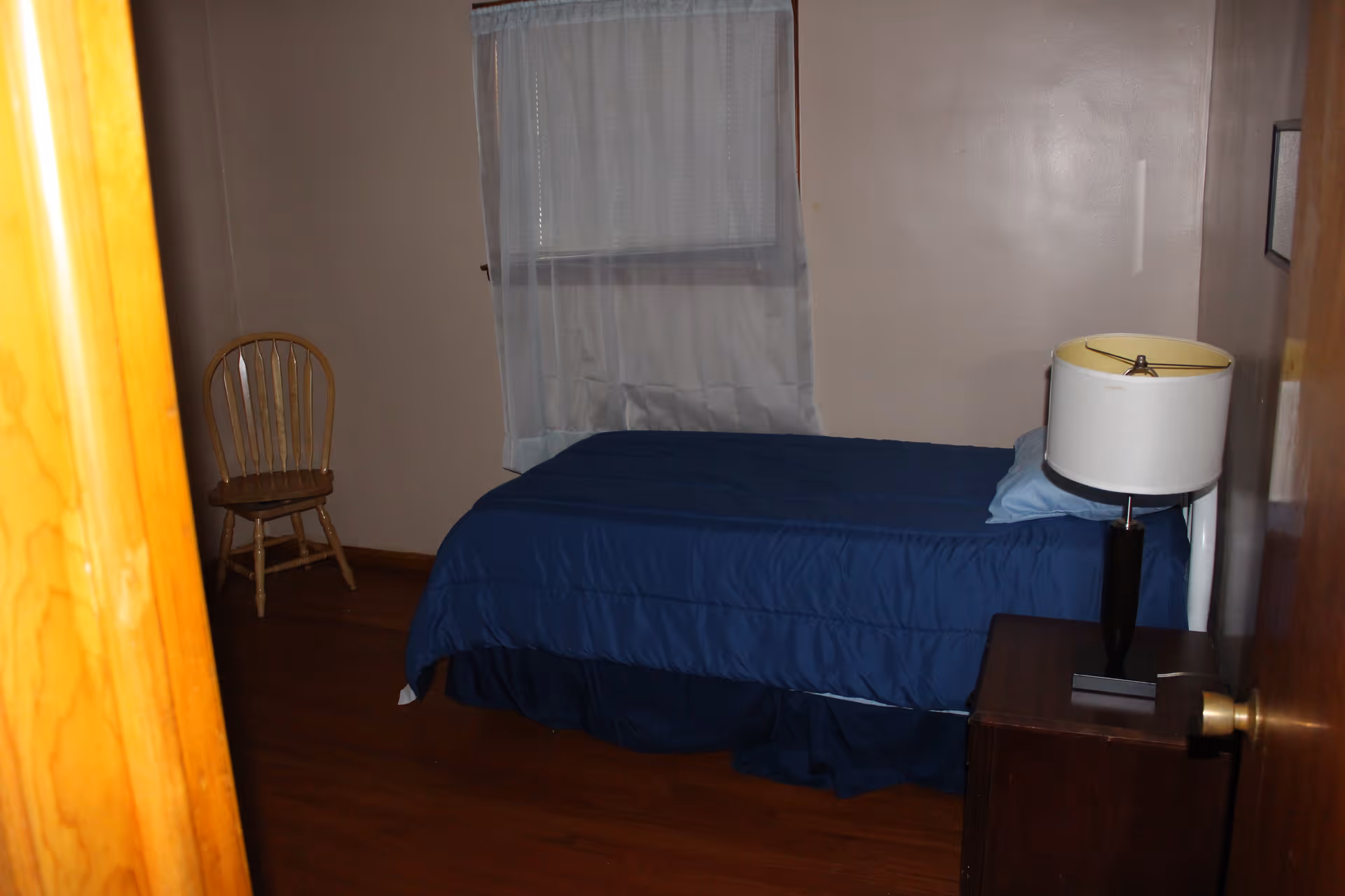 A small bedroom with a single bed covered in a blue comforter and a light blue pillow. There is a wooden chair in the corner near a window with white curtains. A dark wooden nightstand next to the bed holds a lamp with a white lampshade. The room has wooden flooring and beige walls.