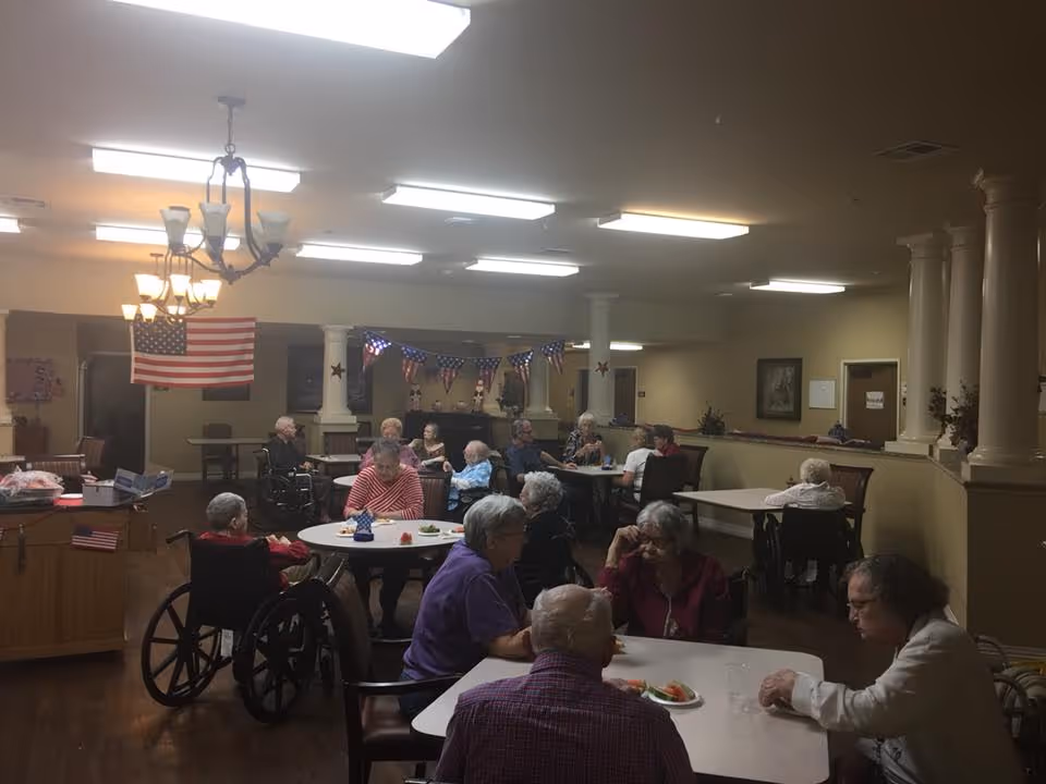 A group of elderly people sitting at tables in a communal dining area of a senior living facility. Some individuals are in wheelchairs, and the room is decorated with American flags and patriotic bunting. The lighting is bright with ceiling fixtures and chandeliers, and the space has columns and beige walls.
