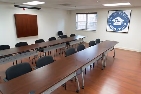 A classroom or meeting room with two long rows of tables and black chairs arranged facing the front. The room has wooden flooring, a window with blinds, a wall-mounted wooden cabinet, and a framed logo on the wall that reads 'RU University 2017'.