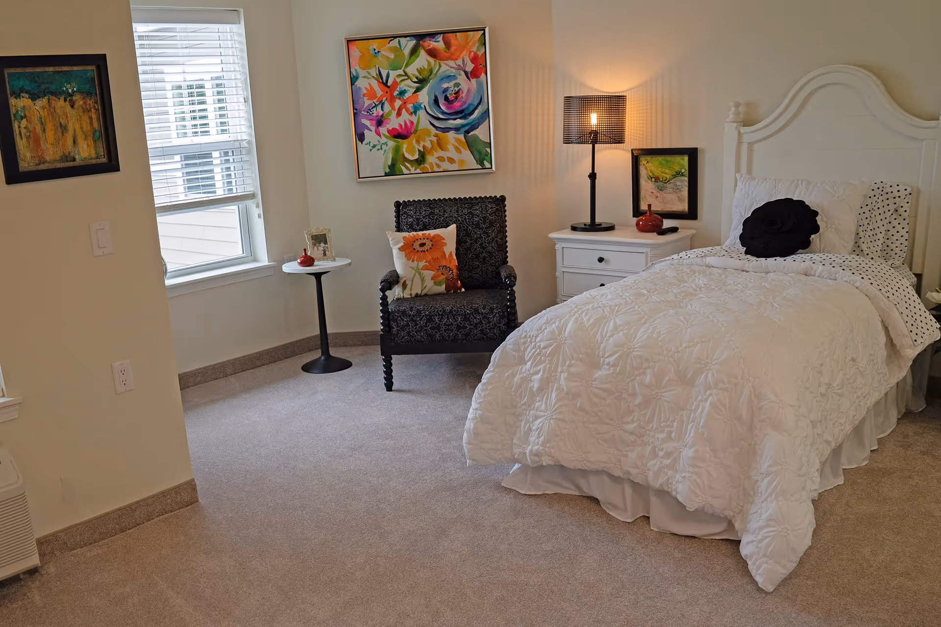 A cozy bedroom with a white bed featuring a textured white comforter and decorative pillows, a white nightstand with a lamp and small artwork above it, a black patterned armchair with a floral pillow, a small round side table with a red vase and picture frame, and two colorful paintings on the walls. The room has beige carpet and a window with white blinds.