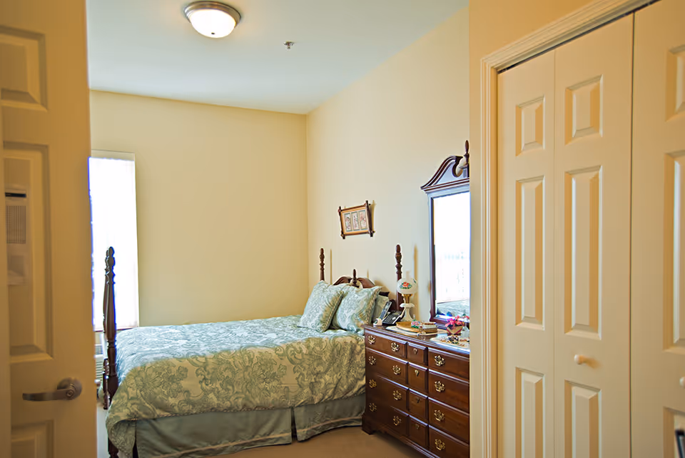 A cozy bedroom with a bed covered in a light green patterned bedspread and matching pillows. There is a wooden dresser with a mirror on the right side of the bed, decorated with a lamp, phone, and small items. The room has beige walls, a ceiling light fixture, and a window with sheer curtains letting in natural light. A partially open door and a closet with double doors are visible in the foreground.