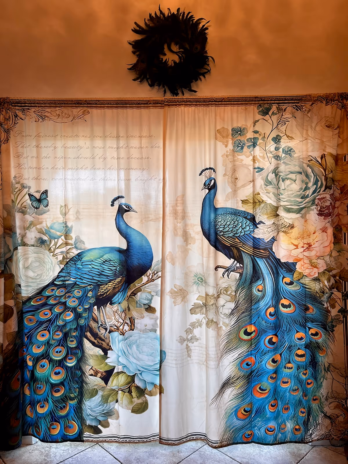 A decorative curtain featuring two large, colorful peacocks surrounded by flowers and a butterfly. Above the curtain is a dark wreath hanging on a warm-toned wall. The curtain has a vintage-style floral and script design.