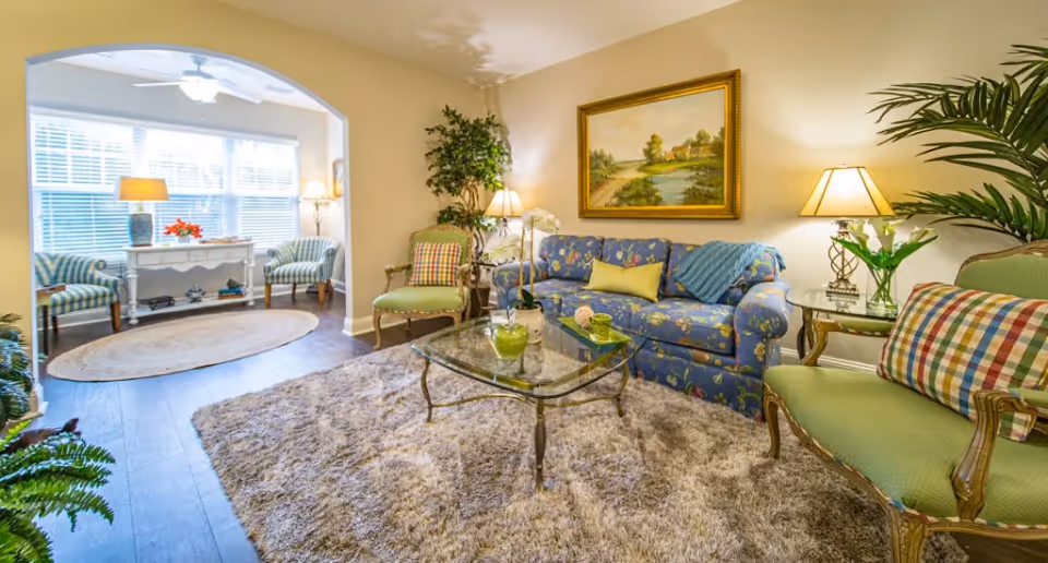 A cozy living room with a blue floral sofa, two green armchairs with plaid cushions, a glass coffee table on a plush beige rug, and a large painting of a landscape on the wall. The room is well-lit with table lamps and natural light from a windowed alcove with two striped chairs and a white console table.