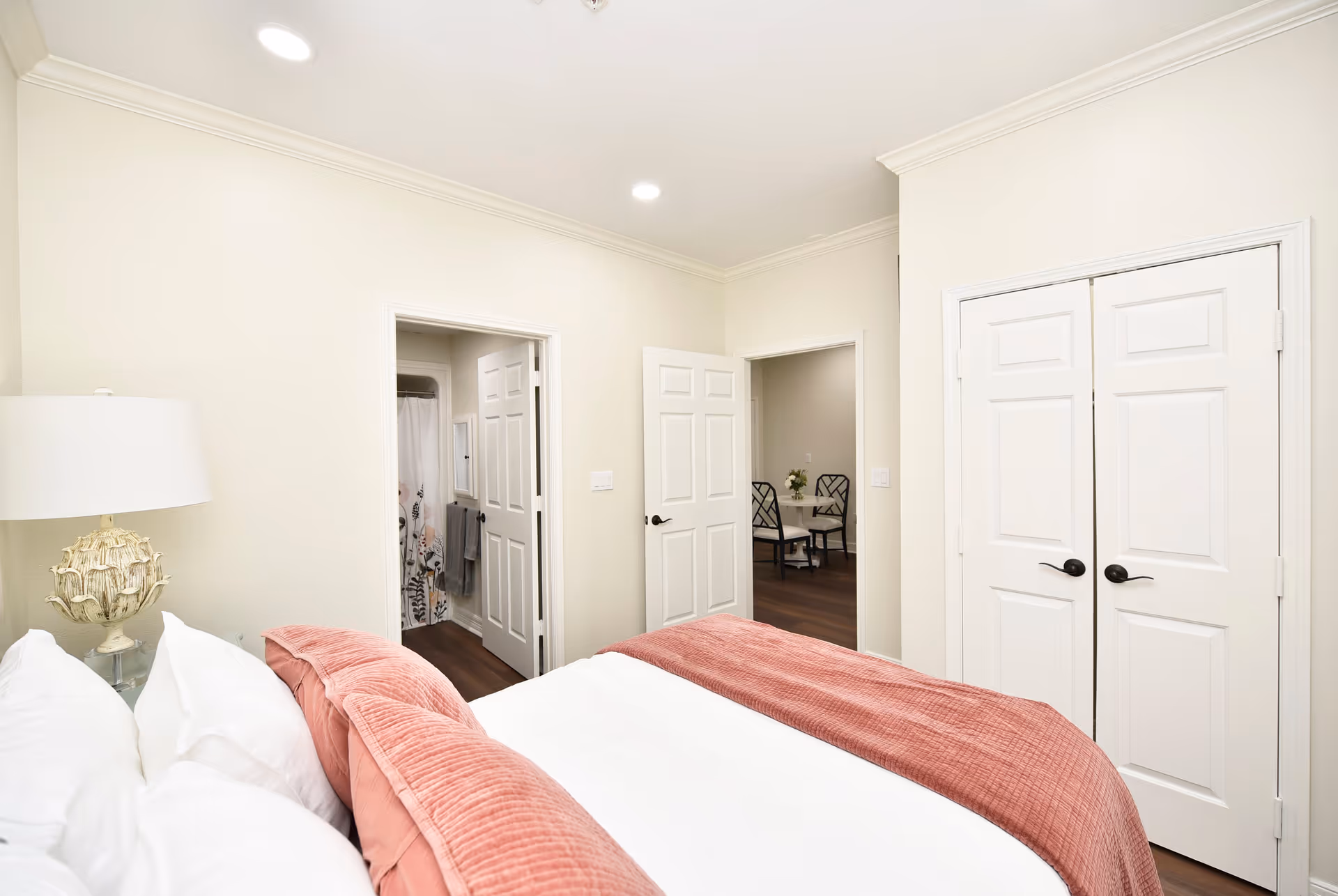 A bedroom with a neatly made bed featuring white sheets and a pink quilted blanket. To the left of the bed is a decorative lamp on a glass nightstand. The room has cream-colored walls and white trim. There are two open doors: one leading to a bathroom with a floral shower curtain, and the other leading to a dining area with a small table and chairs. A closed double-door closet is visible on the right side of the image.