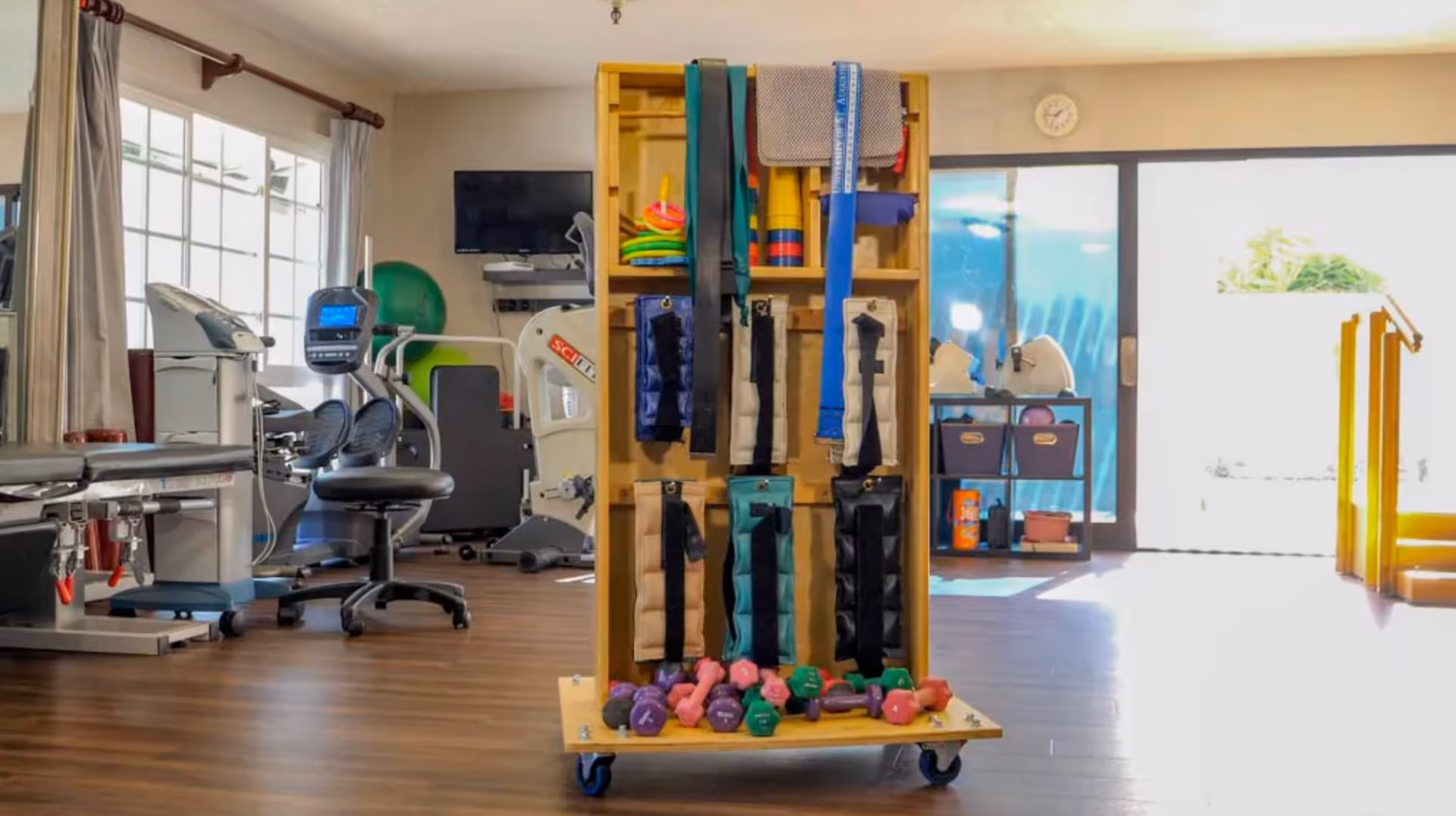 A well-lit physical therapy or exercise room with various fitness equipment including stationary bikes, weights, resistance bands, and exercise balls. The room has large windows with curtains and a door leading outside, allowing natural light to fill the space.