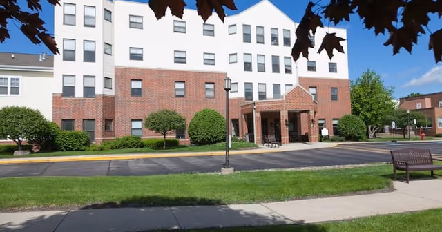Front exterior view of a multi-story senior living facility building with a brick and white facade, a covered entrance, manicured bushes, a bench on the sidewalk, and a clear blue sky with some tree leaves visible at the top.