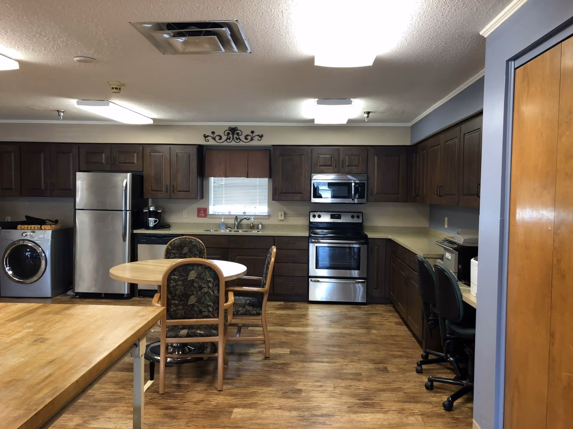 Interior view of a kitchen area in a senior living facility featuring wooden cabinets, a stainless steel refrigerator, dishwasher, stove, and microwave. There is a round wooden table with three chairs in the center, a washing machine on the left, and two office chairs at a counter on the right. The floor is wooden, and the ceiling has fluorescent lighting.