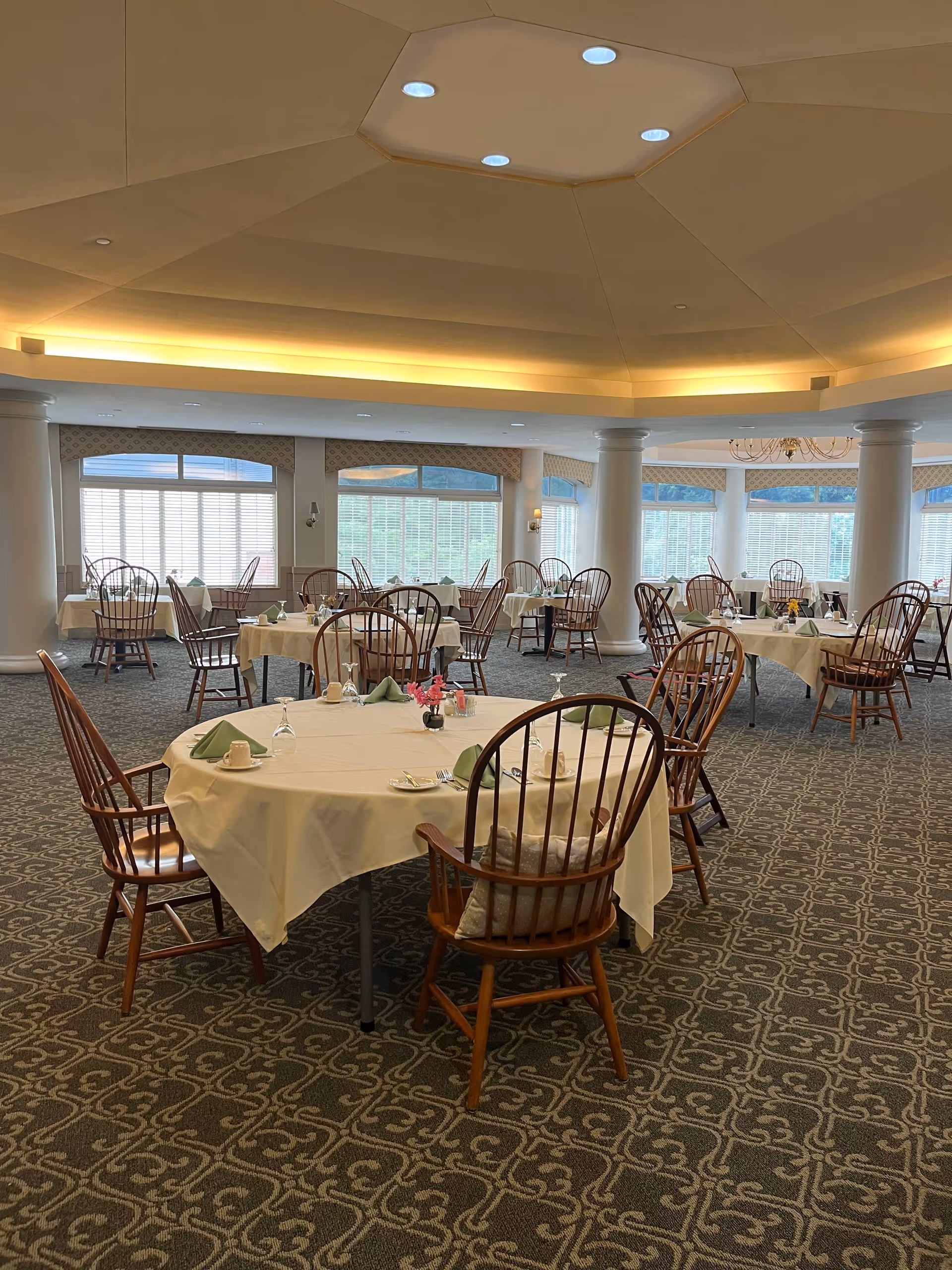 A spacious dining room with round tables covered in white tablecloths, each set with green napkins, cups, and glassware. Wooden chairs with cushions surround the tables. The room features large windows with blinds, white columns, patterned carpet, and a ceiling with recessed lighting.