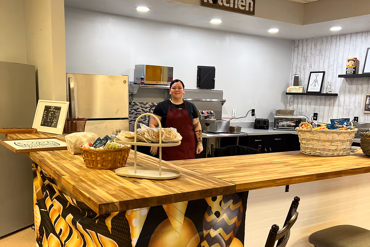 A staff member in a red apron stands behind a wooden kitchen counter with baskets and appliances.