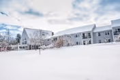 Exterior view of a large senior living facility building covered in snow with a clear sky above and some leafless trees in the foreground.
