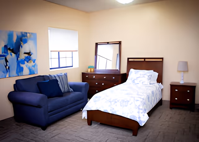 A bedroom with a single bed covered in white and blue patterned bedding, a wooden dresser with a mirror, a wooden nightstand with a lamp, a blue couch with cushions, a window with a white blind, and a blue abstract painting on the wall.