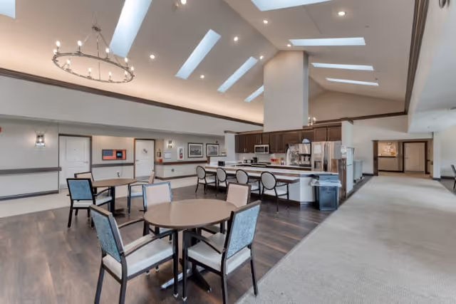 A spacious dining area in a senior living facility featuring round tables with chairs, a kitchen area with a long counter and bar stools, dark wood flooring, and a high ceiling with skylights and recessed lighting.