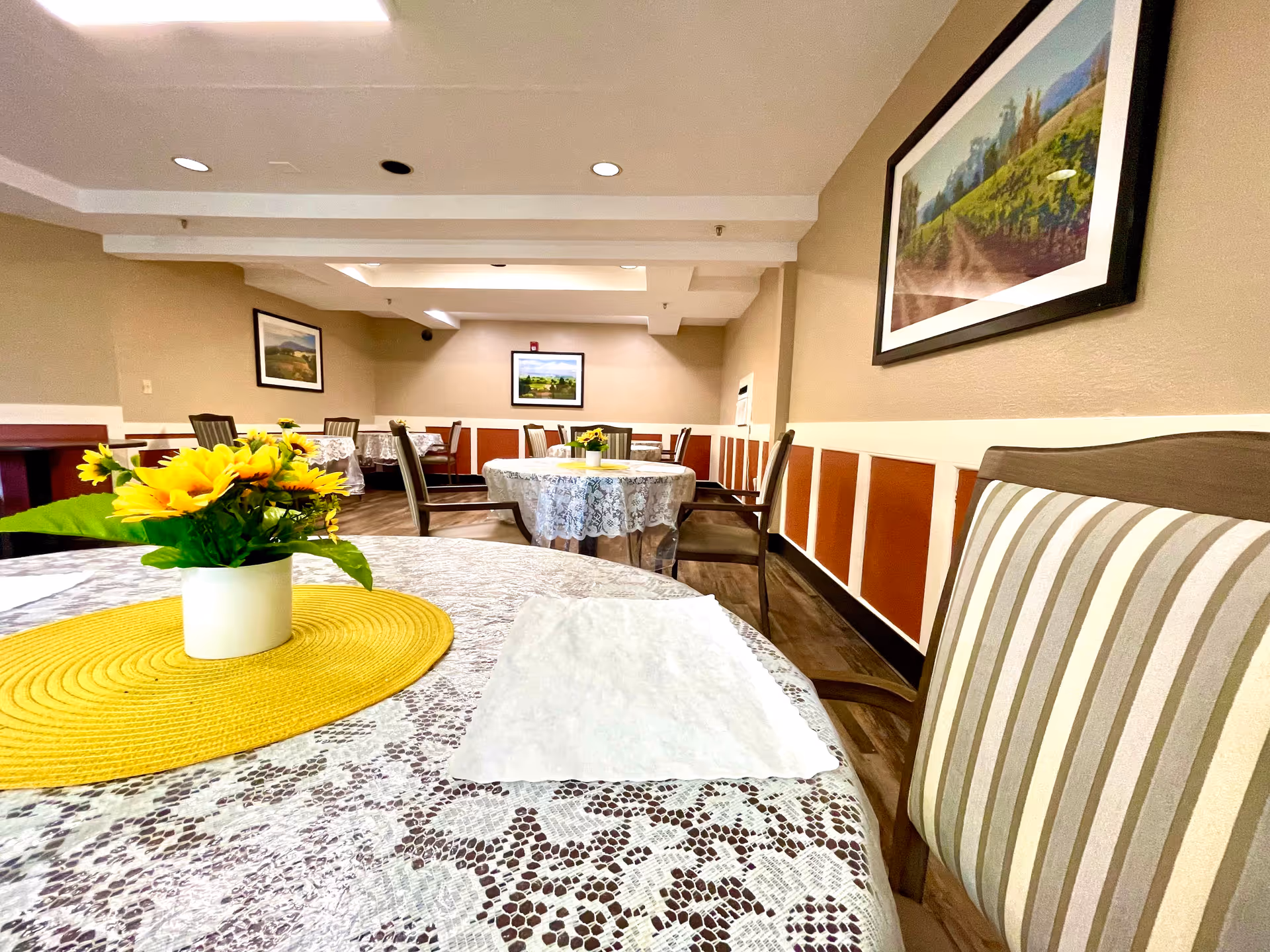 Interior view of a dining room in a senior living facility with round tables covered in lace tablecloths, each adorned with a small vase of yellow flowers on a yellow placemat. The room has wooden chairs with striped cushions, beige walls with framed landscape paintings, and a wooden floor.