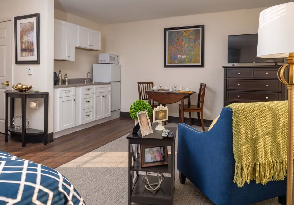 A cozy senior living apartment interior featuring a small kitchenette with white cabinets, a microwave, and a refrigerator. There is a wooden dining table with two chairs set with dishes and glasses. A blue armchair with a yellow throw blanket is positioned near a dark wooden TV stand with a flat-screen TV. A small black side table holds framed photos and a green plant. The room has warm lighting and decorative artwork on the walls.