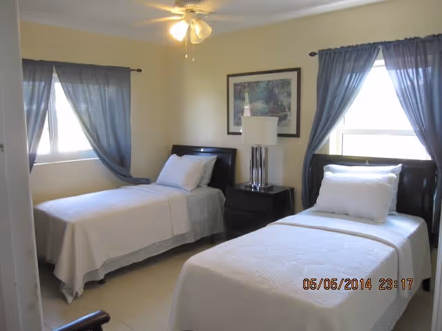 Two twin beds in a bright bedroom with light-colored walls, blue curtains, and a nightstand with a lamp.
