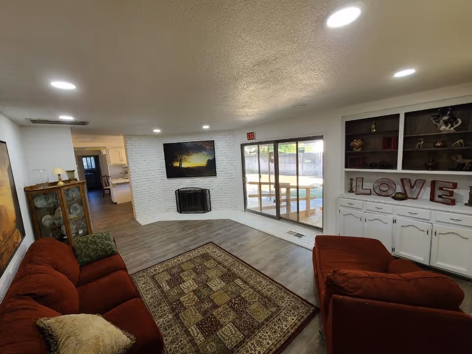 A cozy living room with two red sofas, a patterned area rug, a white brick fireplace with a landscape painting above it, built-in white cabinets and shelves displaying decorative items including letters spelling LOVE, and a glass sliding door leading to an outdoor area with a picnic table.