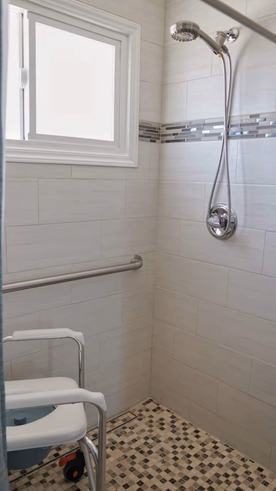 A tiled shower area with a handheld showerhead mounted on the wall, a horizontal grab bar, a window with frosted glass, and a white shower chair with armrests and wheels.