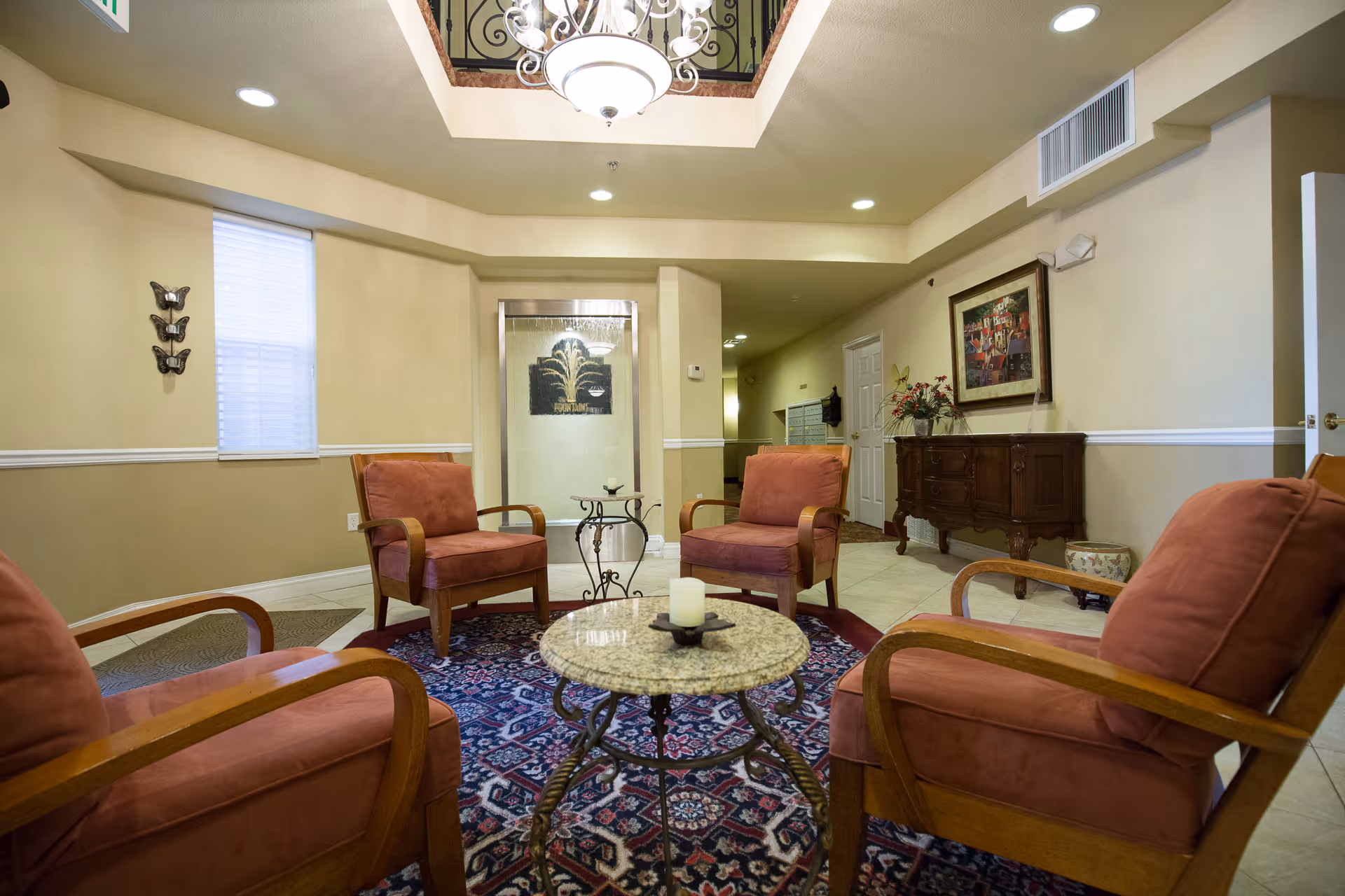 A cozy seating area in a senior living facility with four cushioned armchairs arranged around a round granite coffee table on a patterned rug. The room has beige walls, a window with blinds, decorative wall art, a chandelier hanging from a ceiling opening, and a wooden sideboard with a flower arrangement and framed picture.
