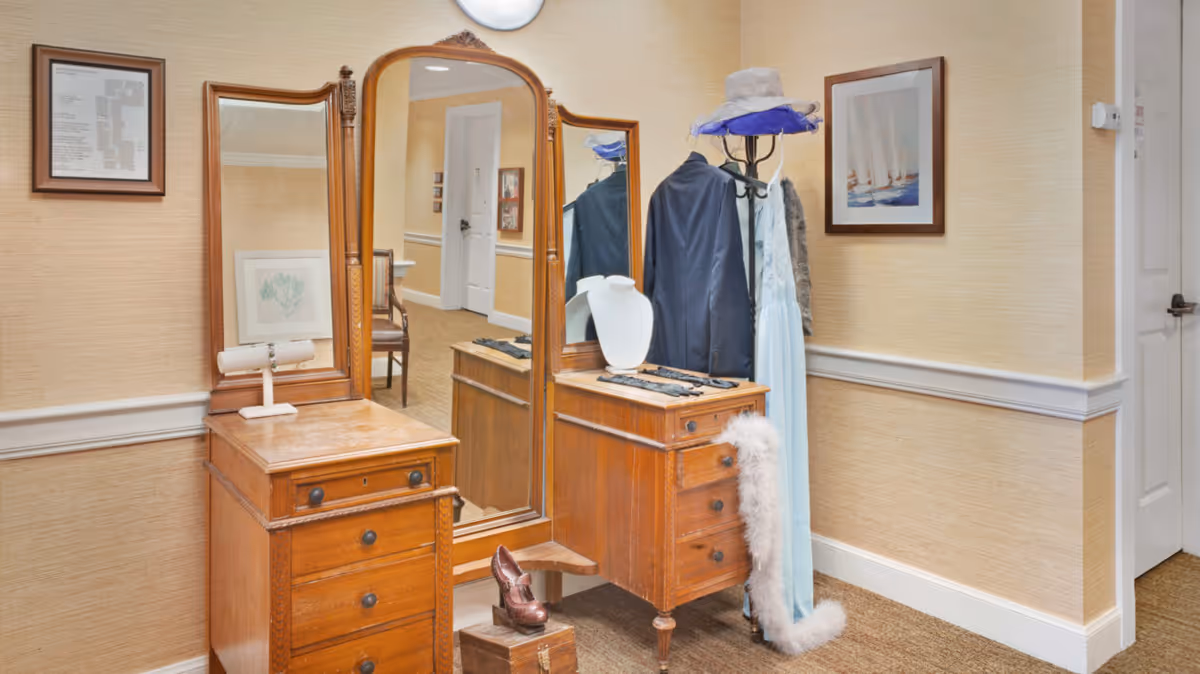 A corner of a room with a vintage wooden vanity dresser featuring three mirrors. On the dresser are a white necklace display bust, gloves, and a feather boa. Next to the dresser is a coat rack holding a dark jacket, a light blue dress, a gray scarf, and a hat with a purple ribbon. The walls are beige with white trim, and there are framed pictures hanging on the walls.