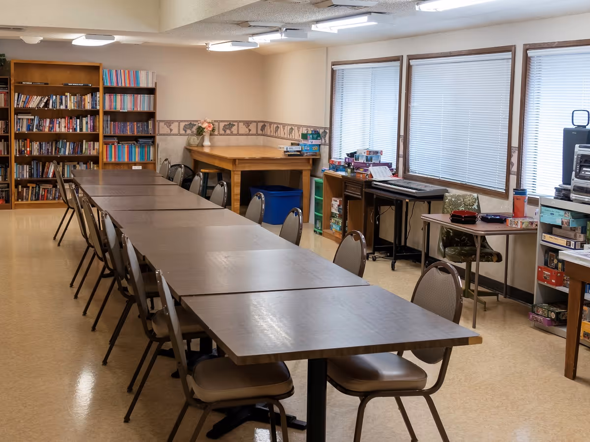 Long multipurpose room with a row of joined tables and chairs, bookshelves, games and large windows along one wall.