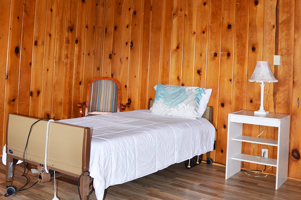 A simple bedroom with wooden paneled walls, featuring a single hospital-style bed with white bedding and a floral pillow, a striped upholstered chair, and a white nightstand with a floral lampshade on top.