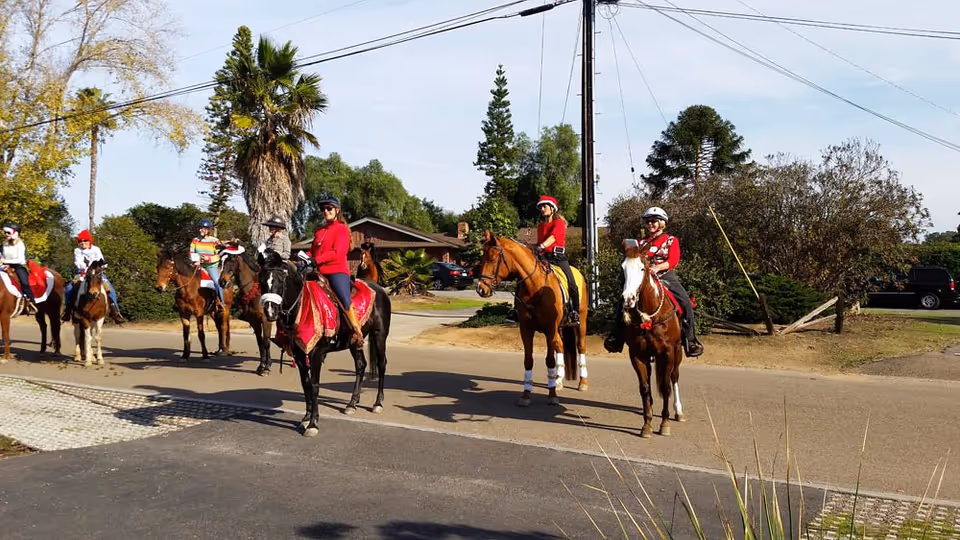 A group of people in festive red attire on horseback paused on a residential street in front of houses and trees.