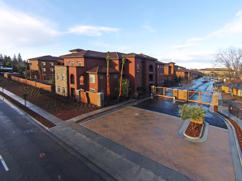 Exterior view of a senior living facility with multiple connected buildings featuring red and brown tones, a gated entrance, landscaped areas with small trees and shrubs, and a paved driveway under a partly cloudy sky.