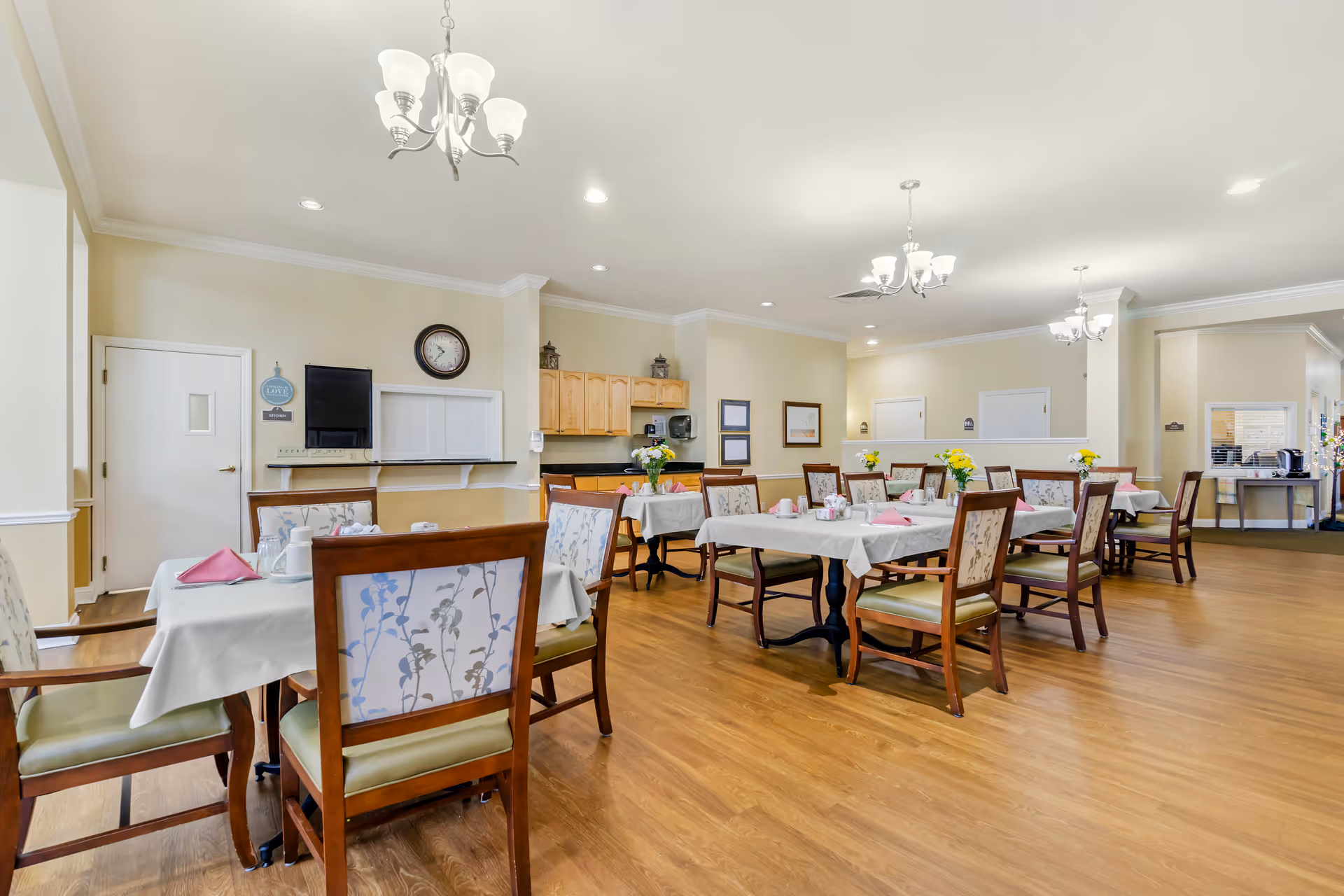 A spacious dining room in a senior living facility with several tables covered in white tablecloths and set with pink napkins, cups, and small flower arrangements. The room has wooden flooring, beige walls, multiple chandeliers, and a small kitchen area with wooden cabinets in the background.