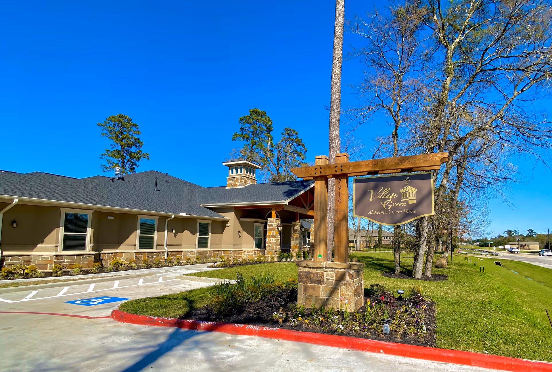 Exterior front of the Village Green memory care building showing its wooden entrance sign, landscaped grounds, and driveway.