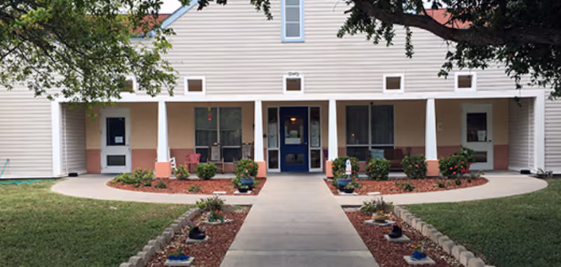 Front exterior view of Villa Northwest Assisted Living building with a concrete walkway leading to a blue door entrance. The building has beige siding with white trim and a covered porch area with columns. There are landscaped garden beds with small bushes and flowers on either side of the walkway, and trees providing shade.