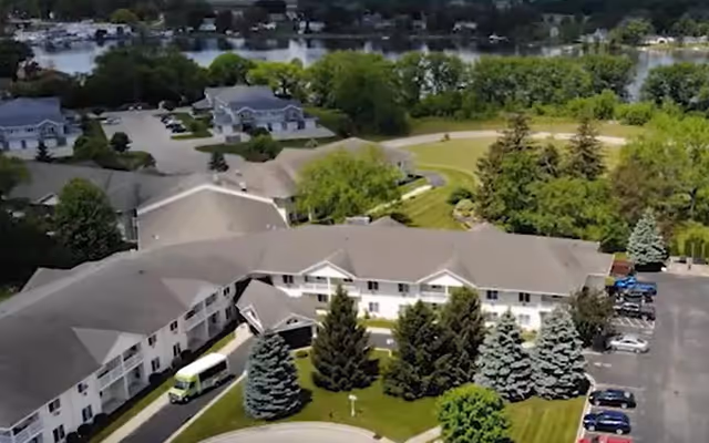 Aerial view of Dimensions Living Beaver Dam facility showing a large two-story building surrounded by trees, a parking lot with several cars, and a green lawn area. In the background, there is a body of water and additional residential buildings.