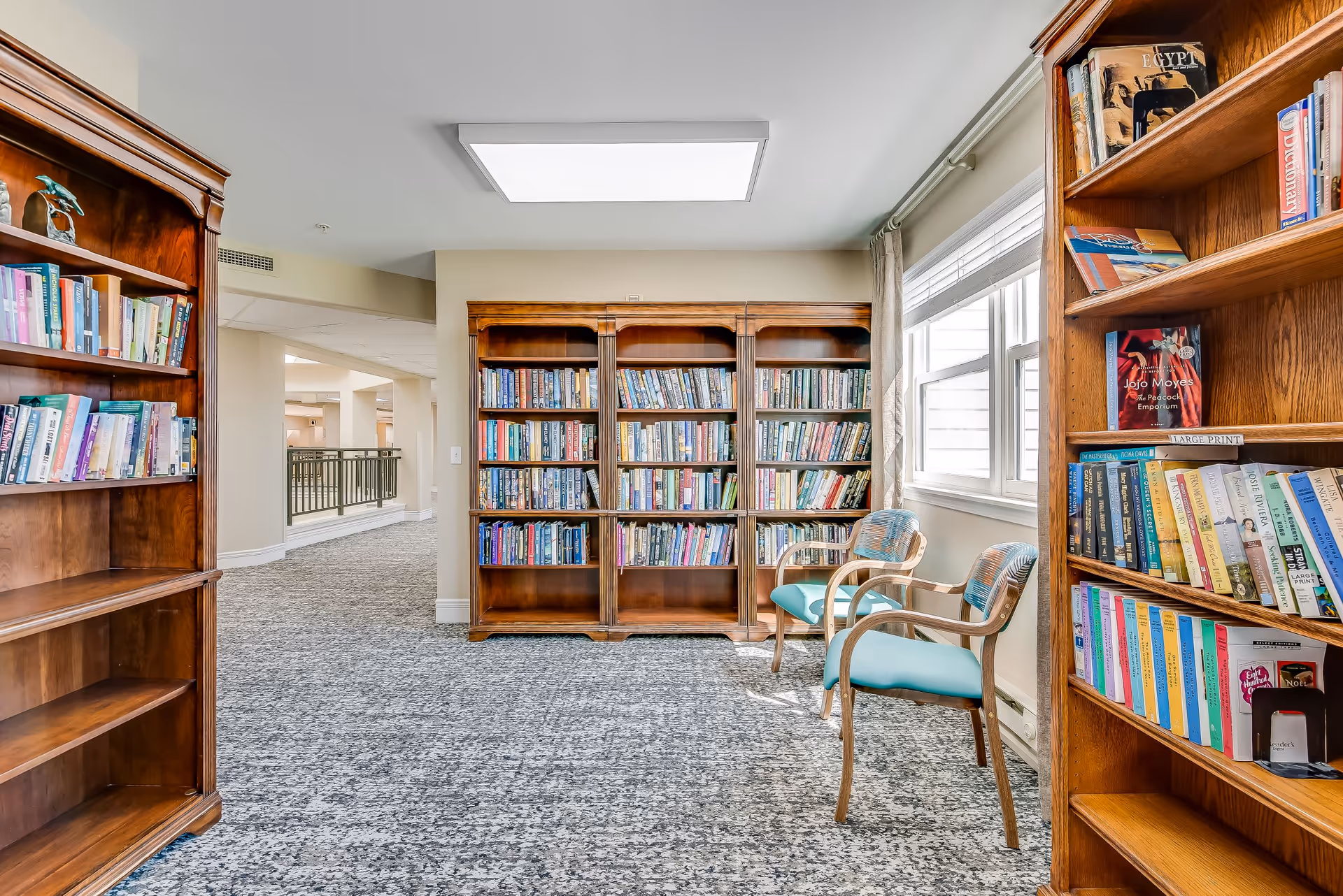 A bright and cozy reading area in a senior living facility featuring three wooden bookshelves filled with books, two wooden chairs with teal cushions, a large window with curtains allowing natural light, and a carpeted floor.