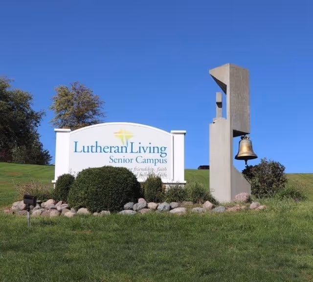 Outdoor view of a sign for Lutheran Living Senior Campus with a bell mounted on a concrete structure next to it, surrounded by grass, bushes, and rocks under a clear blue sky.