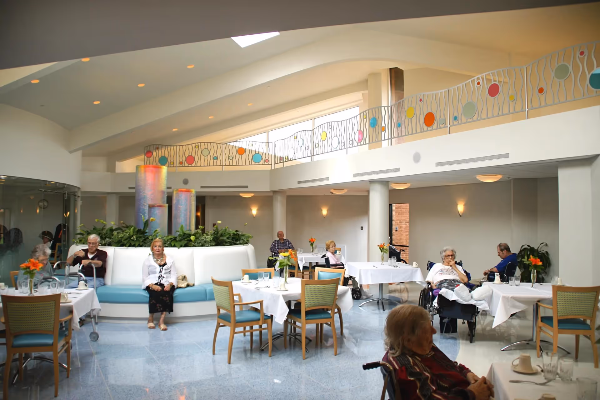 Several elderly residents sit and dine in a bright, open dining room with white tablecloths, a central planter, and a decorative upper-level railing.