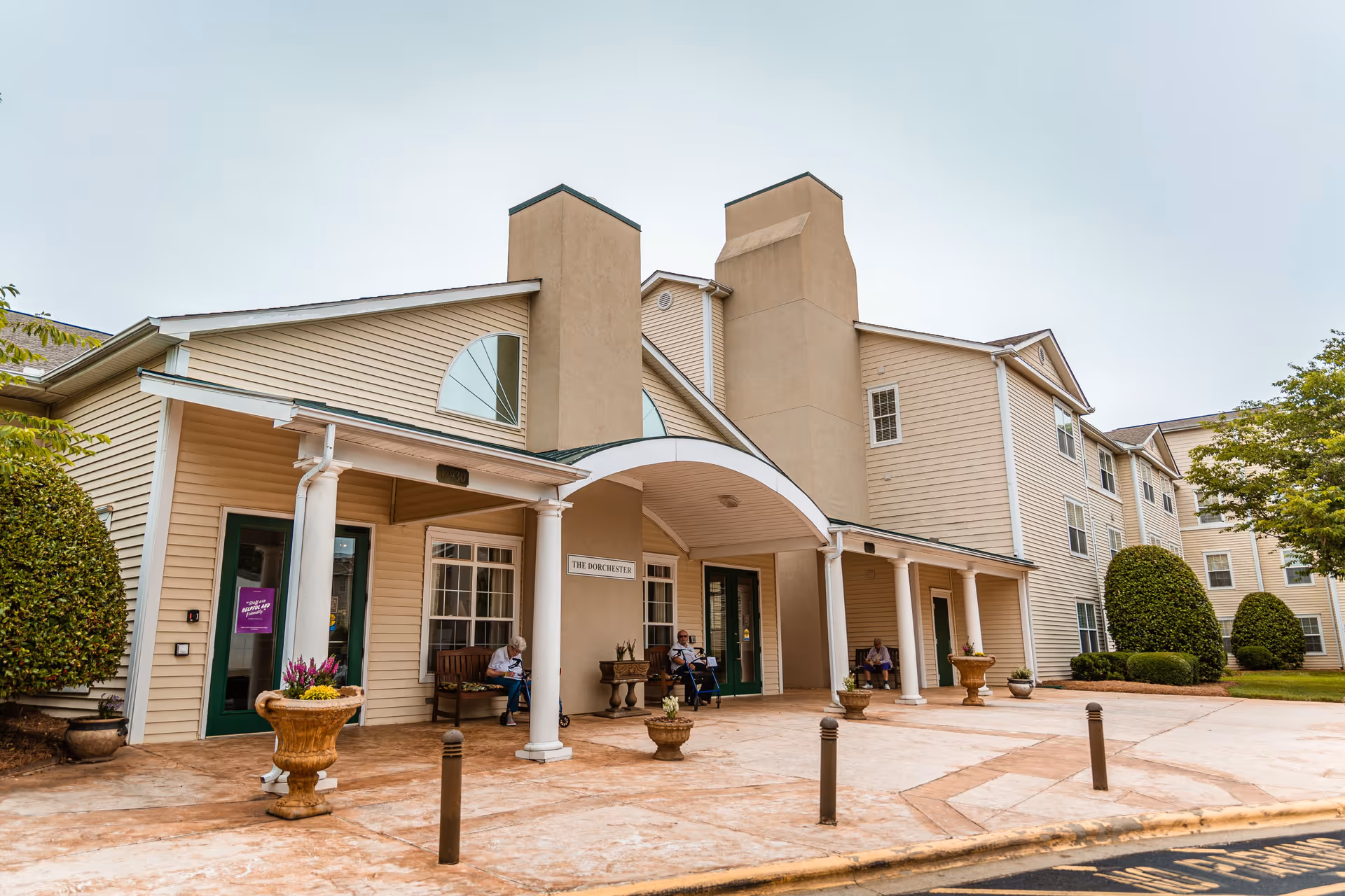 Exterior view of The Dorchester senior living facility showing the entrance with a covered porch supported by white columns. There are three elderly people sitting on benches near the entrance. The building has beige siding and large windows with neatly trimmed bushes and planters with flowers around the entrance area.