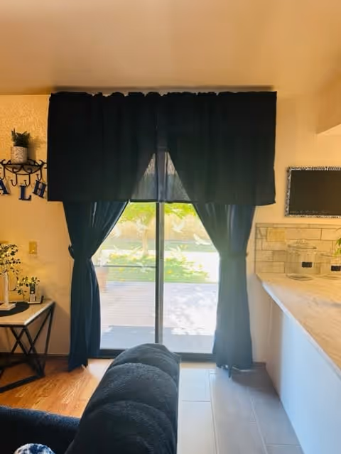 Sliding glass patio door framed by dark curtains with a couch in the foreground and a kitchen counter to the right, showing a view of the backyard.