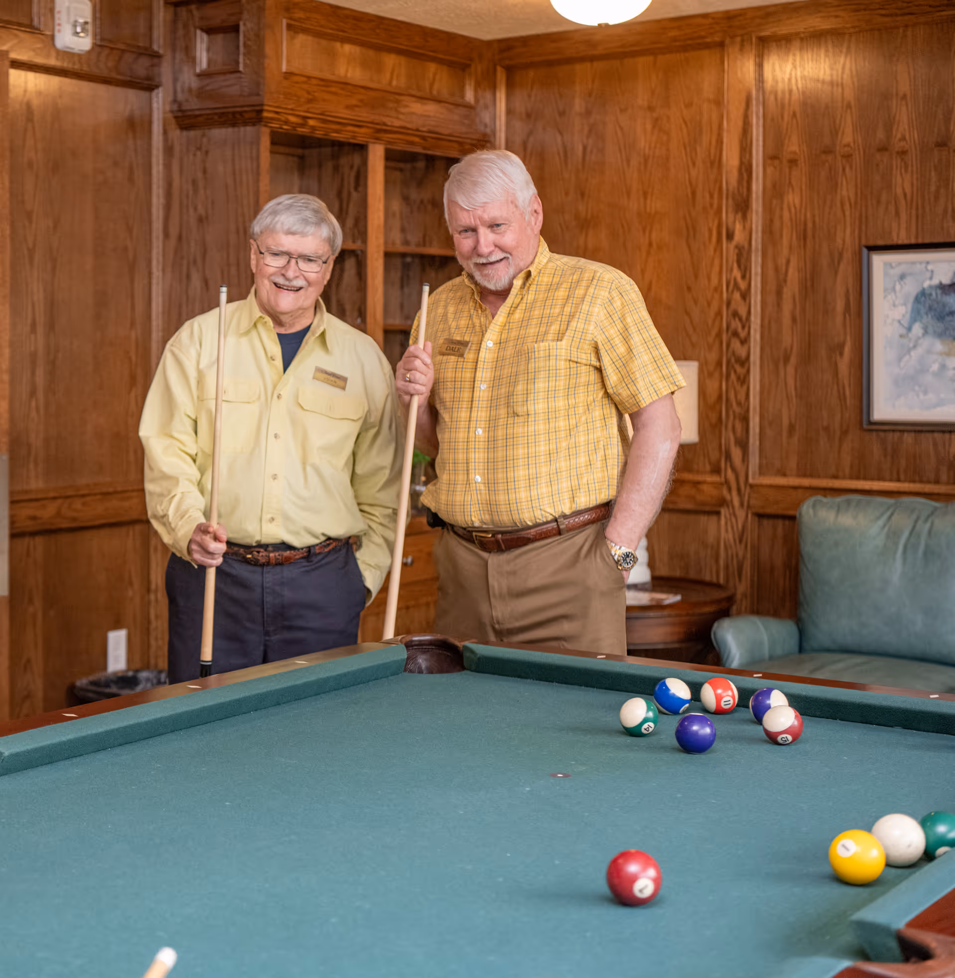 Two elderly men standing next to a pool table in a wood-paneled room, each holding a pool cue and smiling. There is a green couch and a framed picture on the wall in the background.