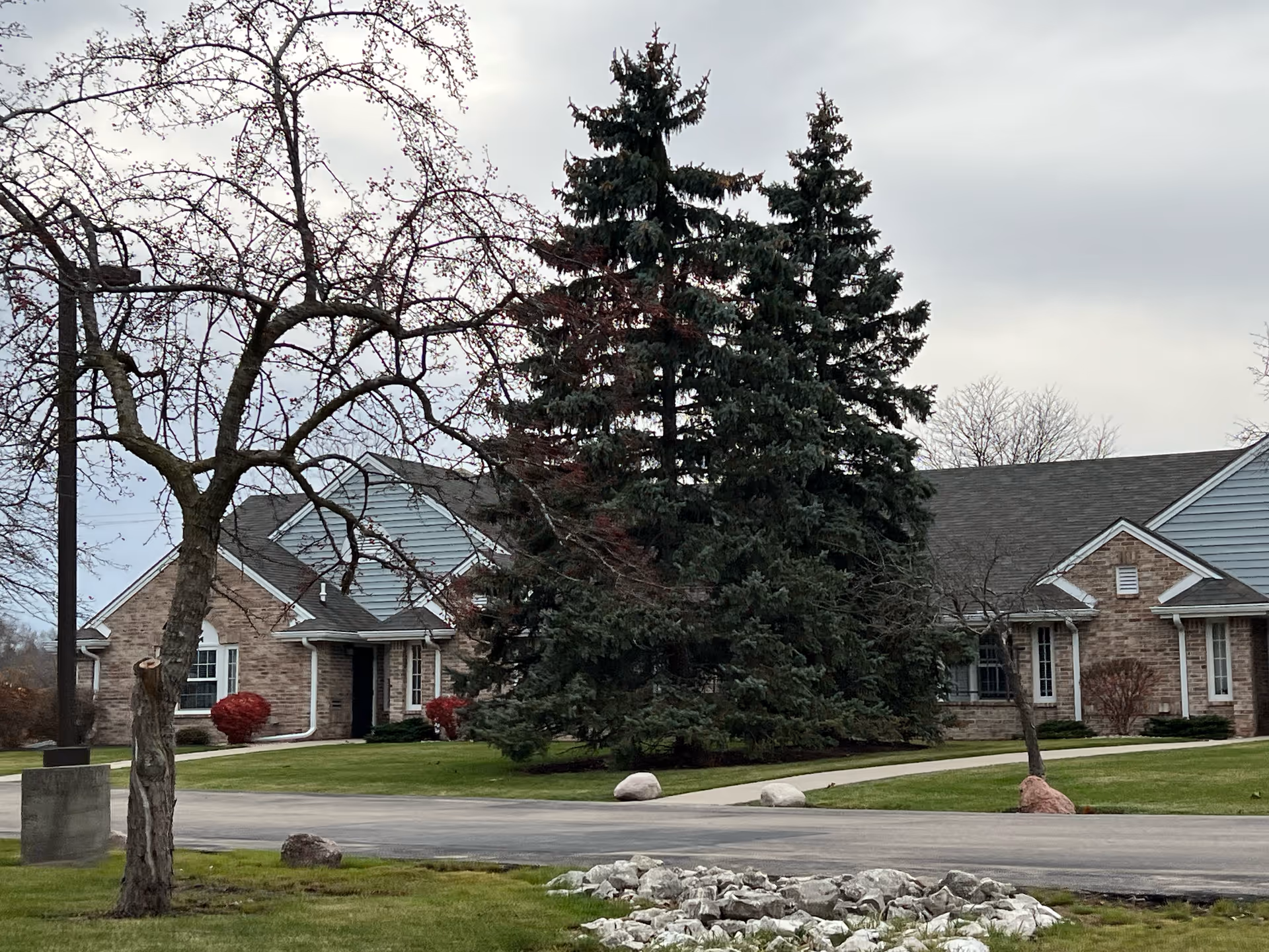 Exterior view of a single-story brick building with a gray roof, surrounded by green grass, leafless trees, and large evergreen trees under a cloudy sky.