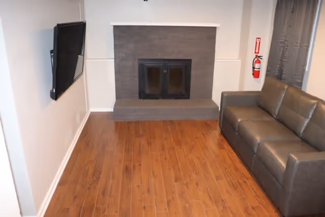 A small living room with wooden flooring, a gray leather couch on the right side, a wall-mounted flat-screen TV on the left, and a gray brick fireplace centered on the back wall. A fire extinguisher is mounted on the wall near the couch.