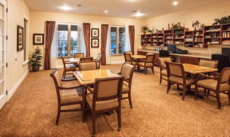 A well-lit room with multiple square wooden tables surrounded by cushioned chairs. The room has large windows with brown curtains, a carpeted floor, and a wall with built-in shelves filled with books and decorative plants. There are also computer workstations along the back wall.