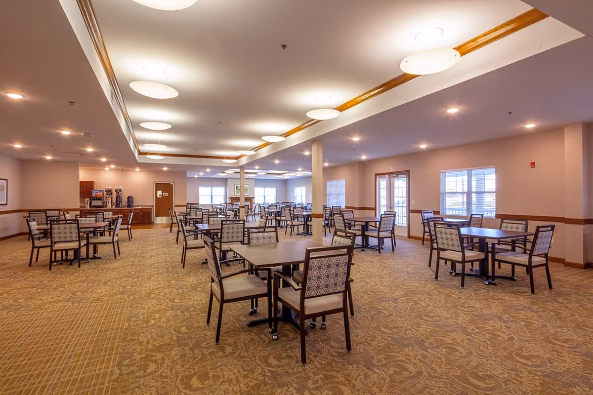 Spacious dining room with multiple square tables and chairs arranged neatly on a patterned carpet. The room is well-lit with ceiling lights and has large windows allowing natural light to enter. There is a beverage station with coffee machines and dispensers against one wall.