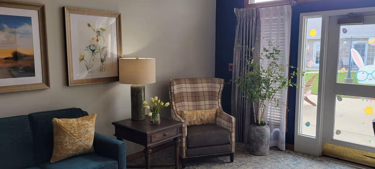 A cozy living room area with a teal sofa and a plaid armchair, each with a decorative pillow. Between them is a wooden side table with a lamp and a small vase of flowers. On the wall above the sofa are two framed pictures. To the right, there is a large window and a glass door decorated with colorful Easter-themed stickers, including a bunny with glasses. A tall potted plant is placed near the window.
